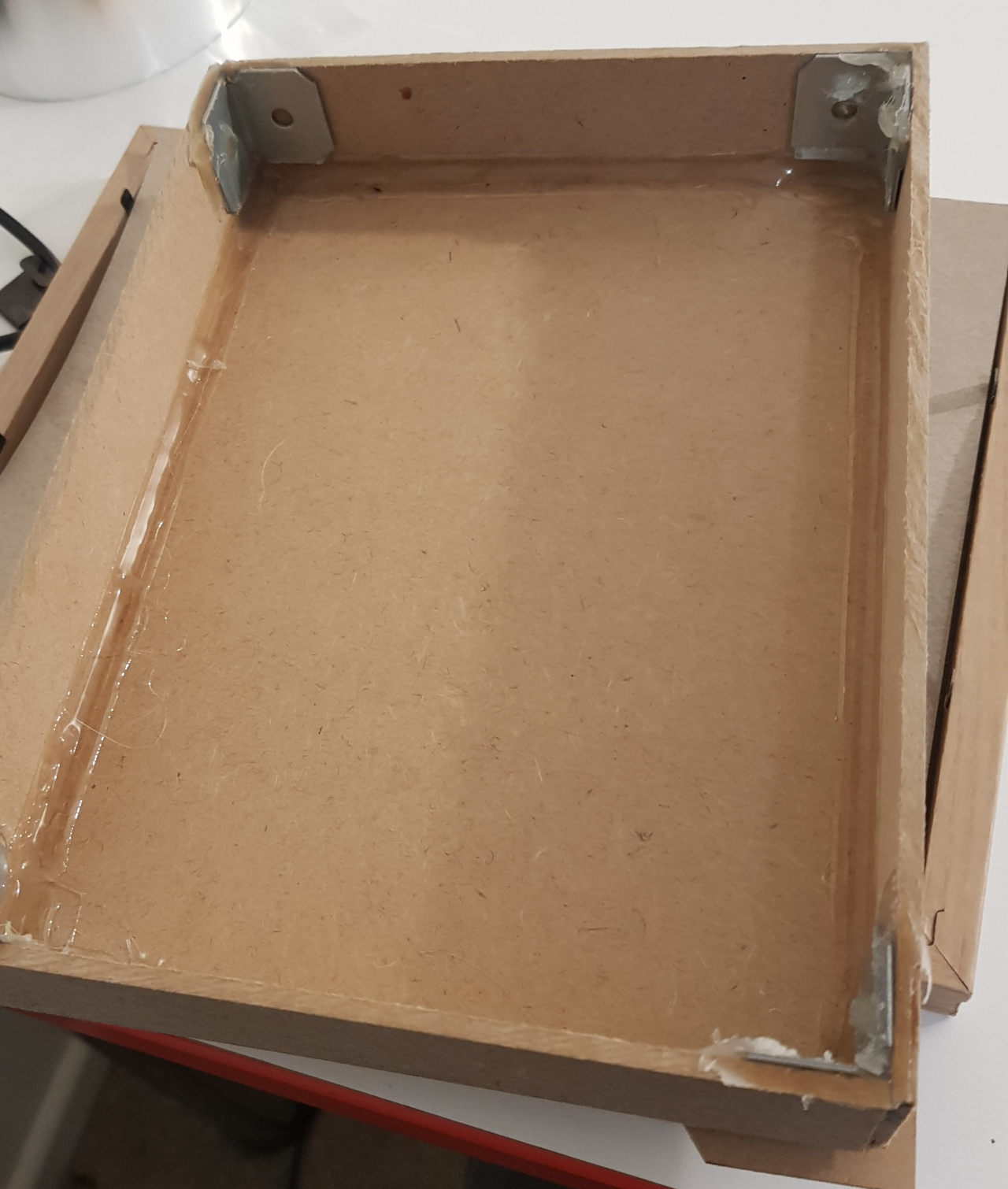 The inside of a fabricated MDF box, showing angle brackets at the corners affixed with hot glue, and a back panel affixed with a bead of hot glue along the inside edge.